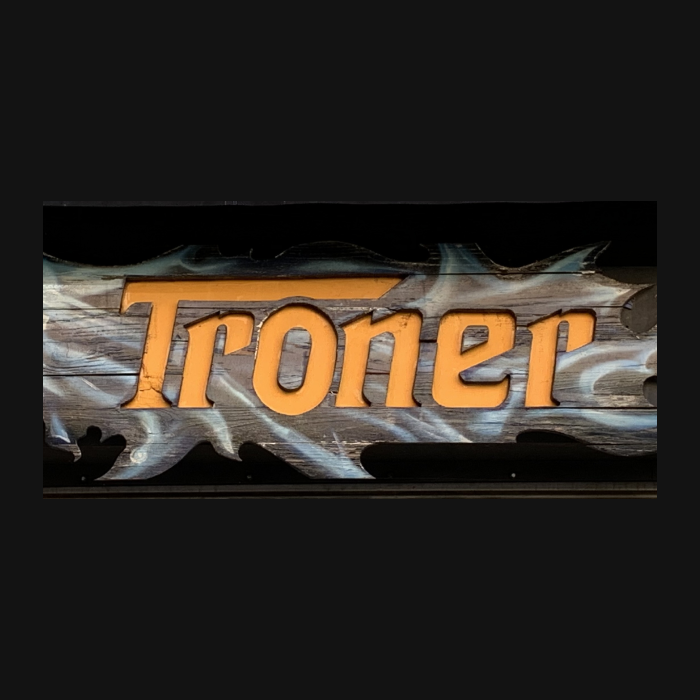 La imagen muestra un letrero con el nombre "Troner" en una tipografía gruesa y estilizada de color naranja. El texto está superpuesto sobre lo que parece ser una representación artística de una tabla de madera o un listón alargado con bordes irregulares y una textura que simula la madera desgastada. En el extremo izquierdo del letrero, se observa una ilustración de una cabeza de dragón o criatura fantástica con detalles en tonos de verde, amarillo y blanco. La criatura tiene un ojo grande y prominente, y su lengua se extiende hacia el principio de la letra "T" de la palabra "Troner".  El estilo general del letrero es rústico y aventurero, evocando un tema de fantasía o medieval que podría estar asociado con un bar temático, una tienda de juegos, o un evento relacionado con juegos de rol o historias de fantasía. El uso del dragón como elemento gráfico sugiere un enfoque en lo mítico o legendario.