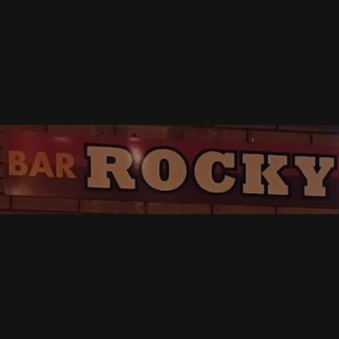 La imagen muestra un rótulo con el nombre "Bar Rocky", destacado en grandes letras corpóreas montadas sobre una superficie que parece una pared de ladrillo oscuro. Las letras son de color rojo oscuro con bordes dorados, lo que les da un efecto tridimensional y llamativo. El nombre "ROCKY" está en una tipografía audaz y alargada, y el término "BAR" está situado encima en un tamaño más pequeño. El diseño tiene un estilo clásico y robusto, sugiriendo un ambiente acogedor y temático, posiblemente relacionado con música rock o cine, lo cual podría ser atractivo para los visitantes que aprecian estos temas.